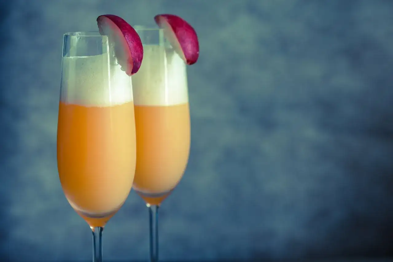 Dwa kieliszki z pianką i owocowym napojem, ozdobione plasterkami brzoskwini. Czy to Mimosa czy Bellini?