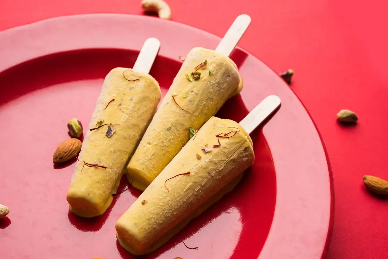 Trzy lody kulfi na patyku, ozdobione szafranem i orzechami, na czerwonym talerzu. Idealny przepis na kulfi!