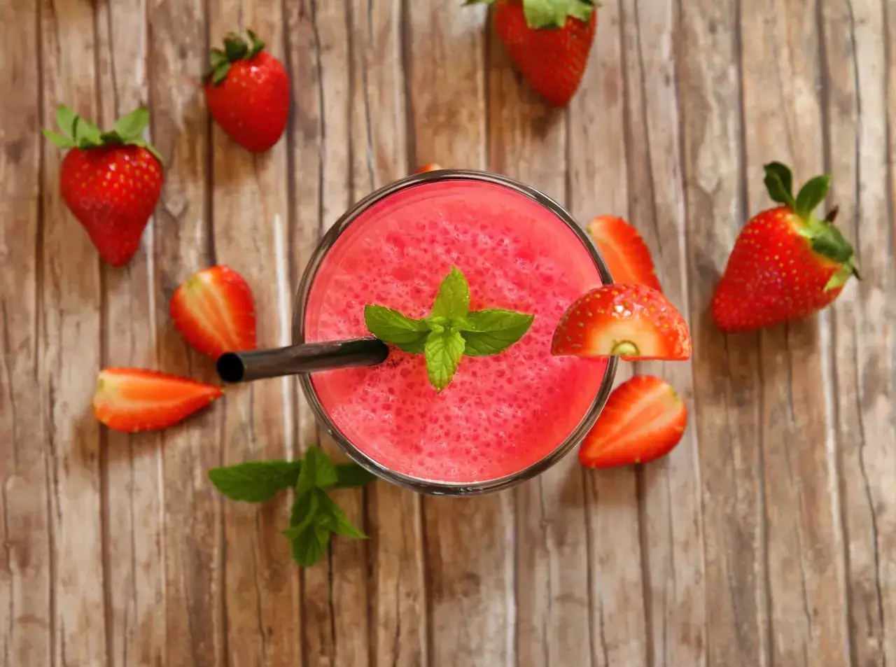 Orzeźwiający drink z truskawkami, ozdobiony miętą, otoczony świeżymi owocami na drewnianym tle.