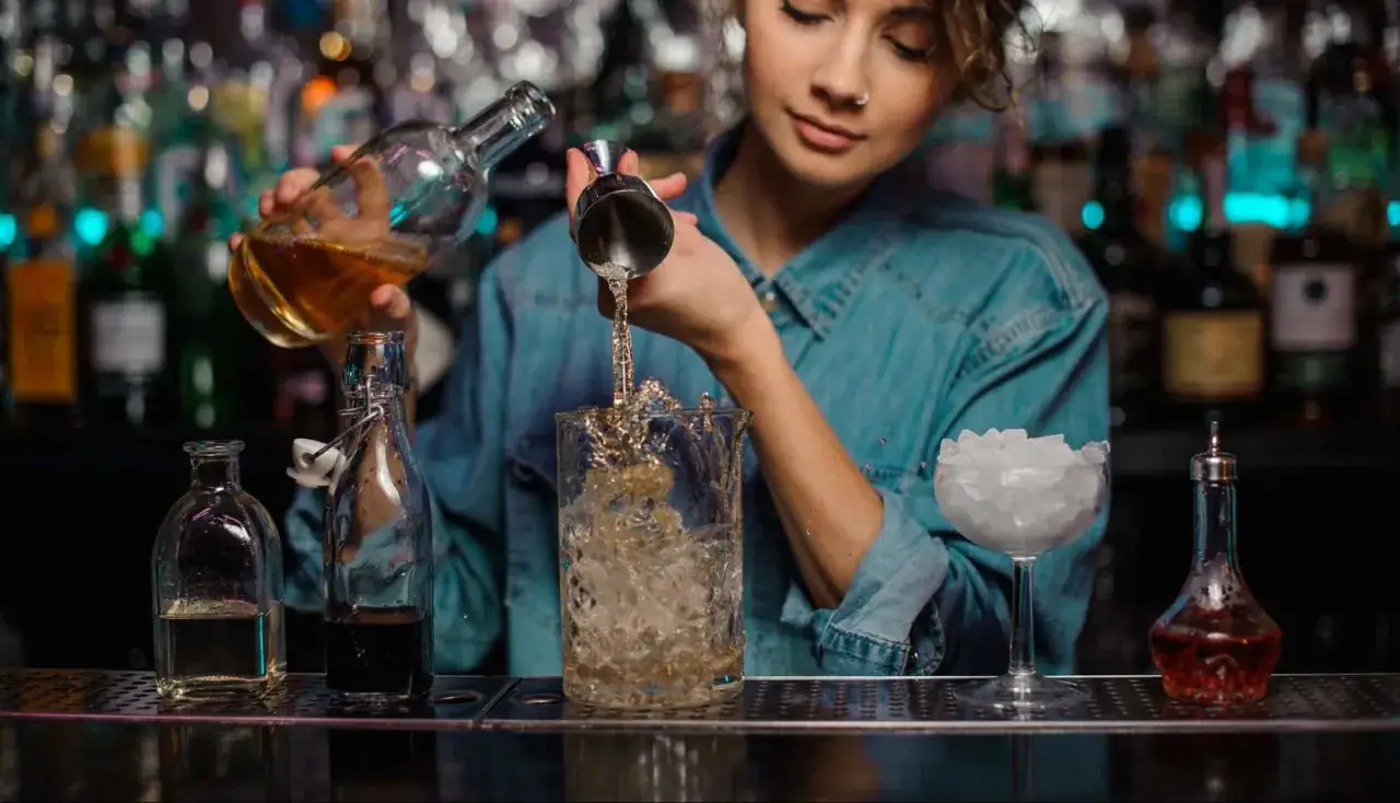 Młoda barmanka przygotowuje drinka, przelewając płyn z butelki do shakera. To początek drogi, jak zostać barmanem.